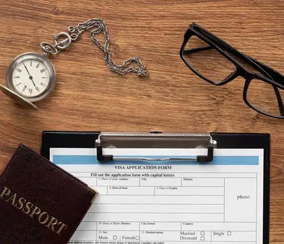A visa application form on a clipboard is placed on a wooden desk, alongside a pair of black glasses, a pocket watch with a chain, and a partially visible passport.
